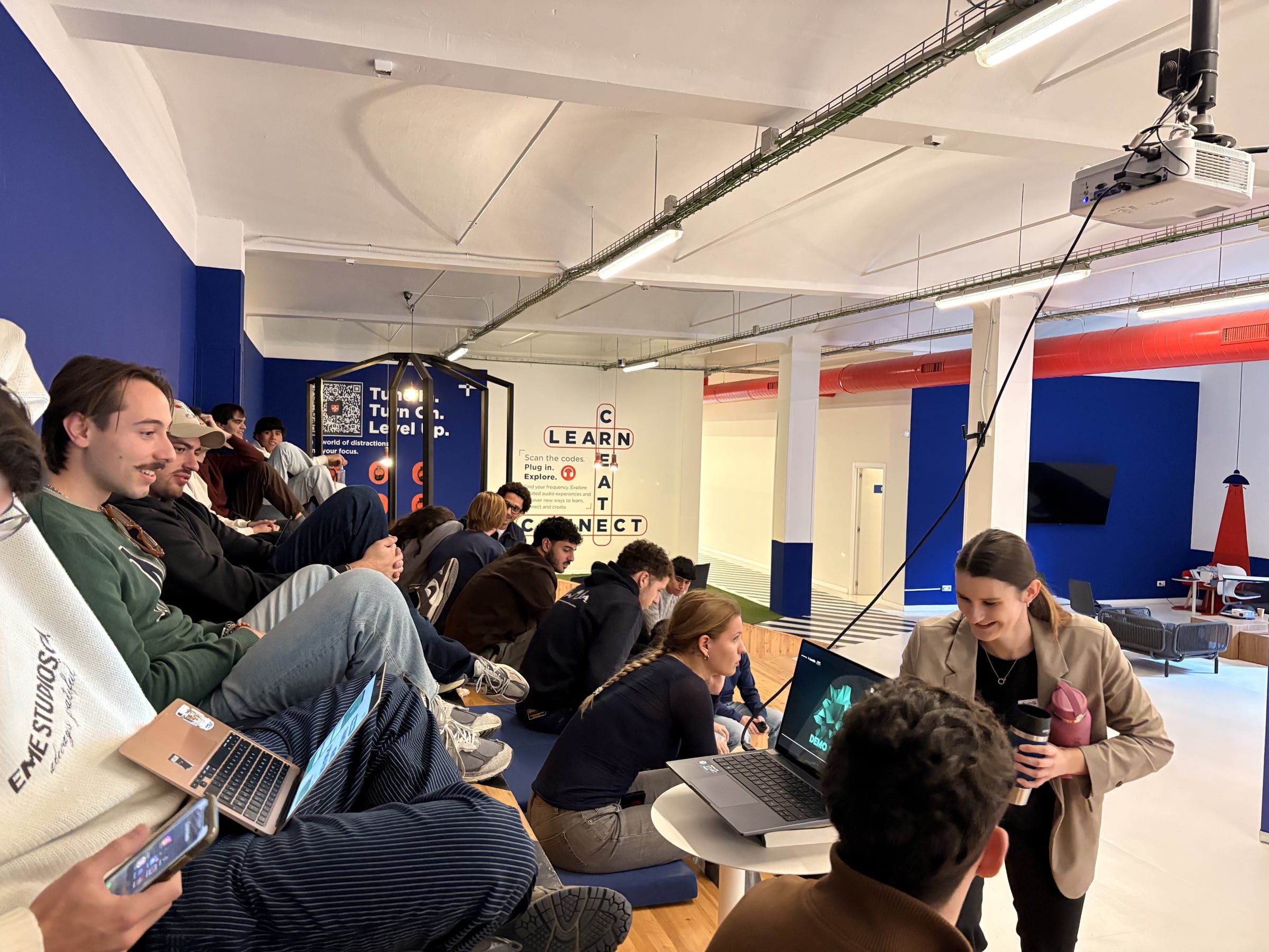 Students on cushions and benches with laptops open during BCN Hack 2025, one team showing a demo