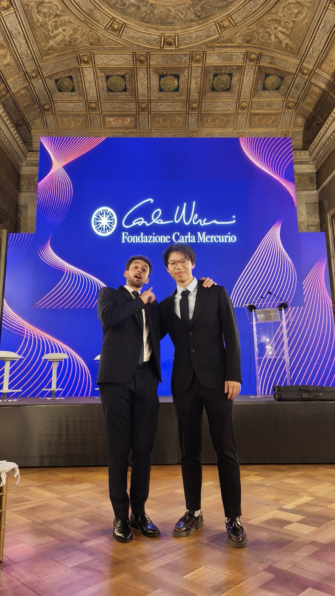 Two people in suits on stage in front of a purple 'Fondazione Carlo Mercurio' screen