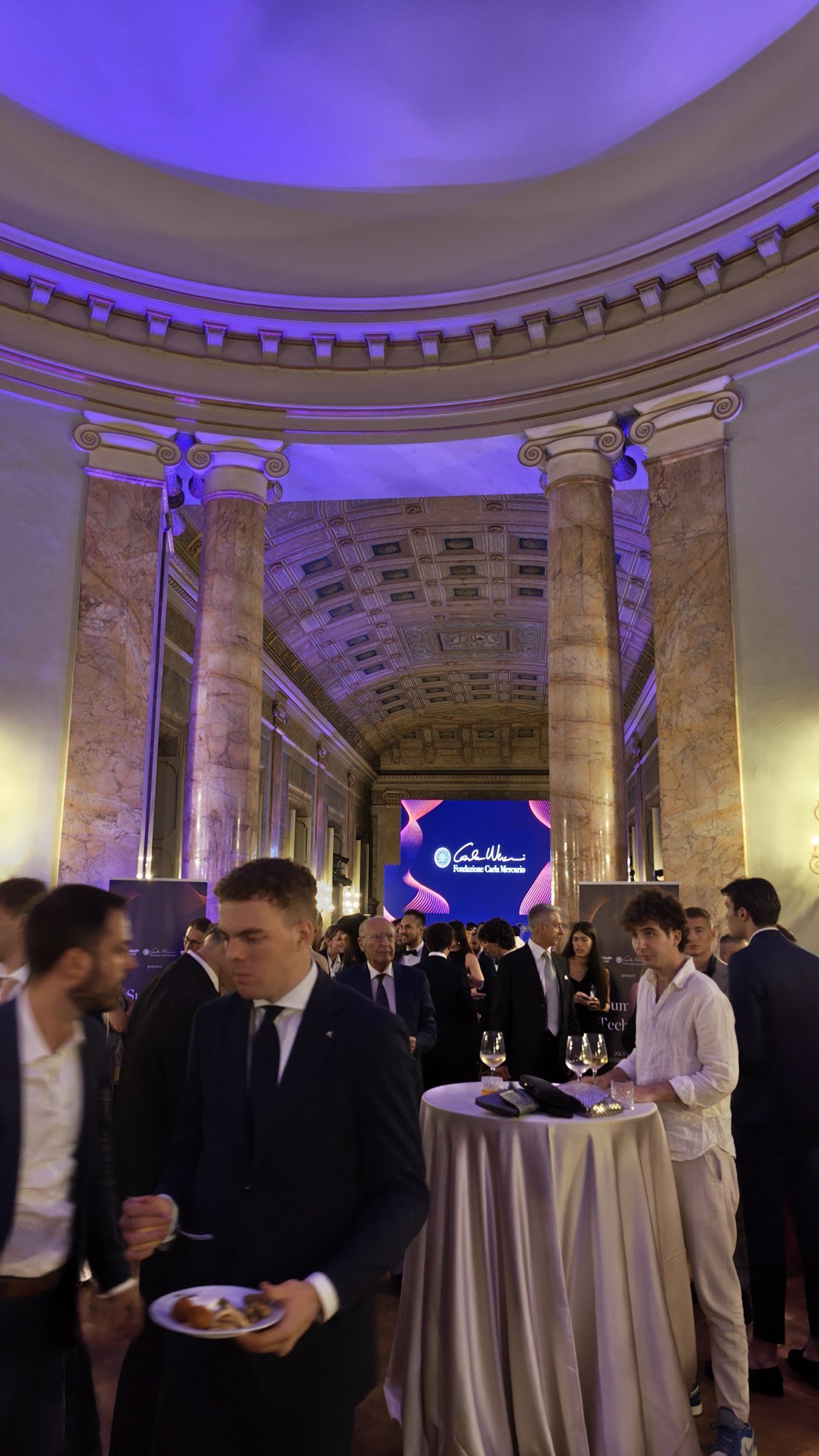 Guests in suits and dresses talking around high tables under a domed, purple-lit ceiling