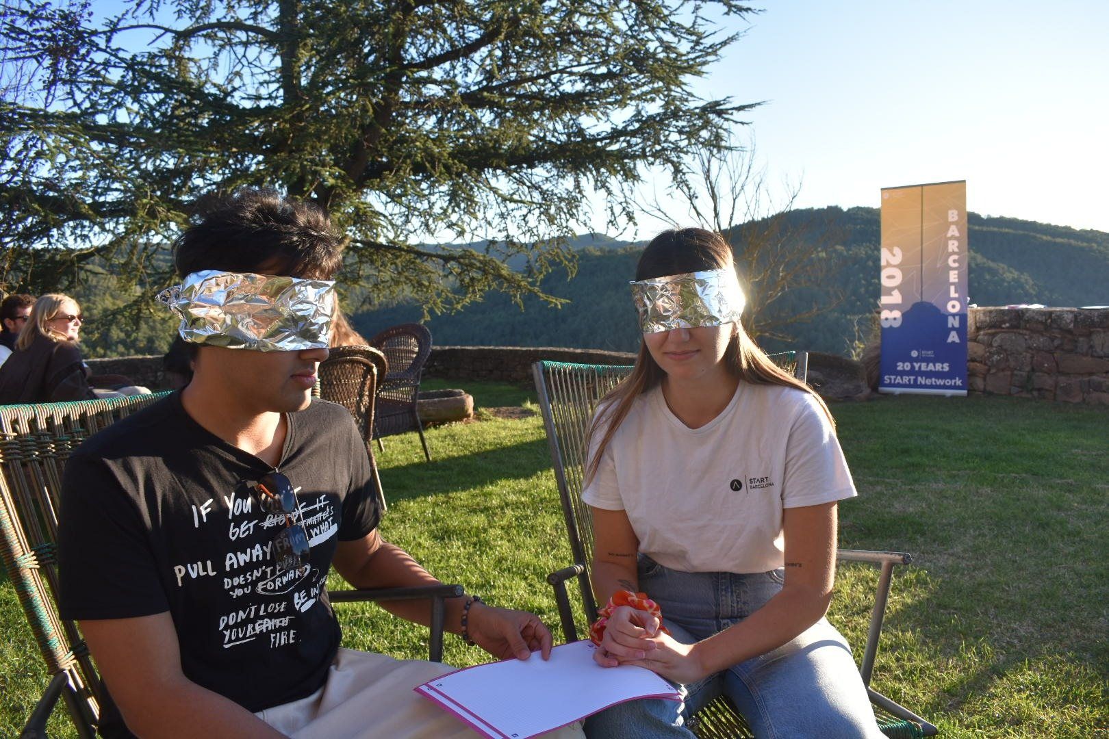 Two members in garden chairs wearing tinfoil blindfolds during a team exercise, hills behind them