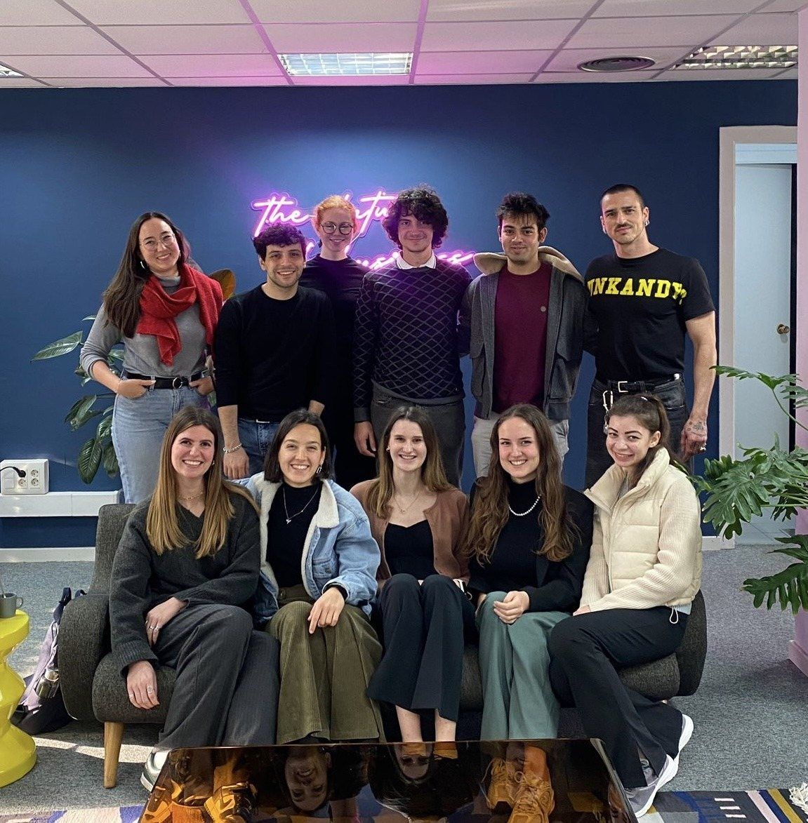 Group photo of members and hosts on and around a sofa in an office with a pink neon sign
