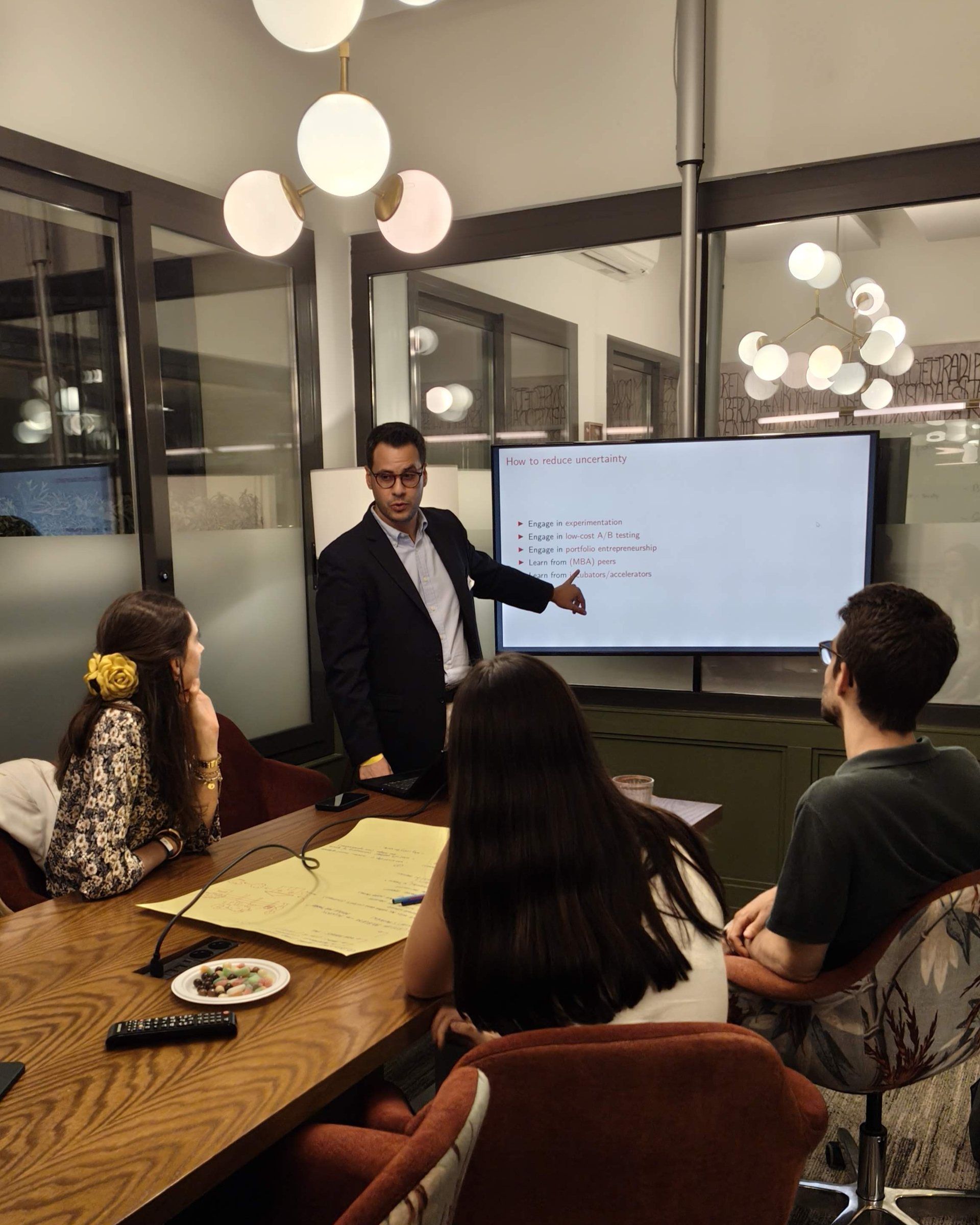 A mentor in a blazer walking a team through a slide in a meeting room, notes and snacks on the table