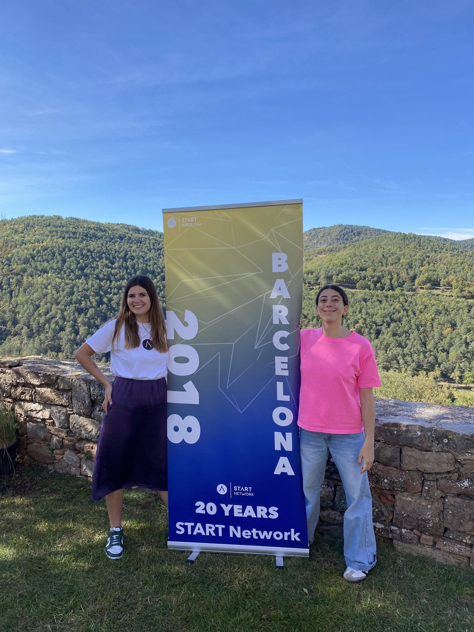 Two members either side of the START Barcelona banner on a terrace above wooded hills