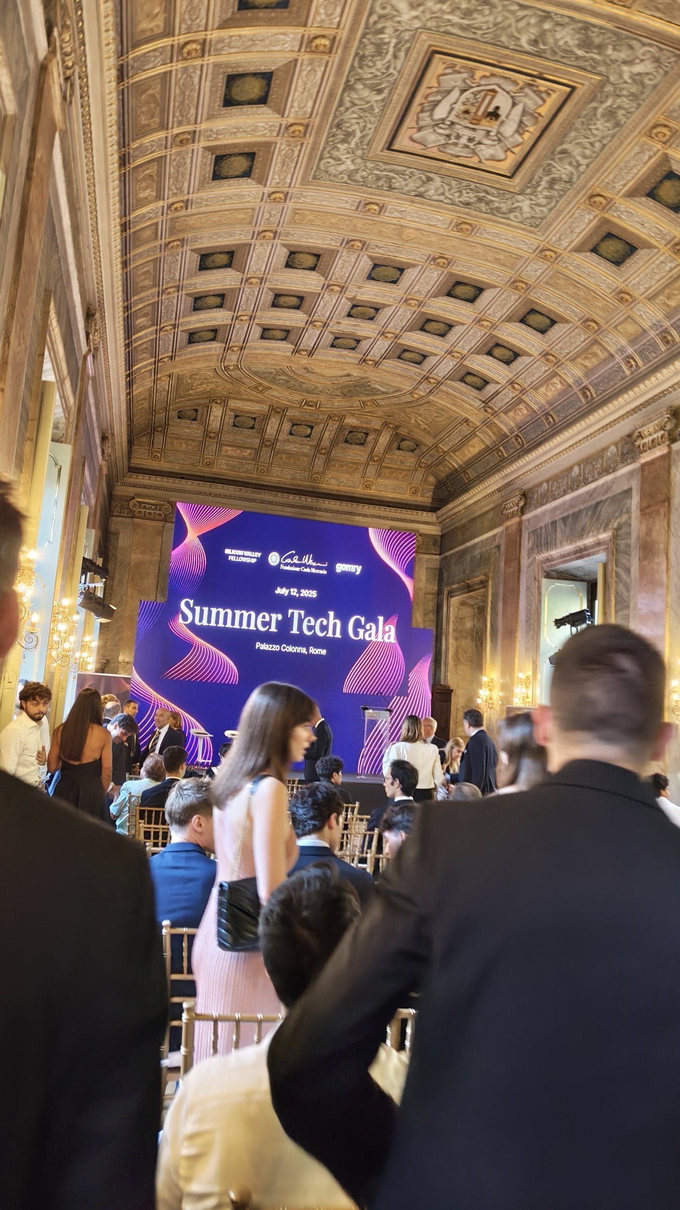 Guests in a gold-ceilinged hall at the Summer Tech Gala, the event screen at the far end