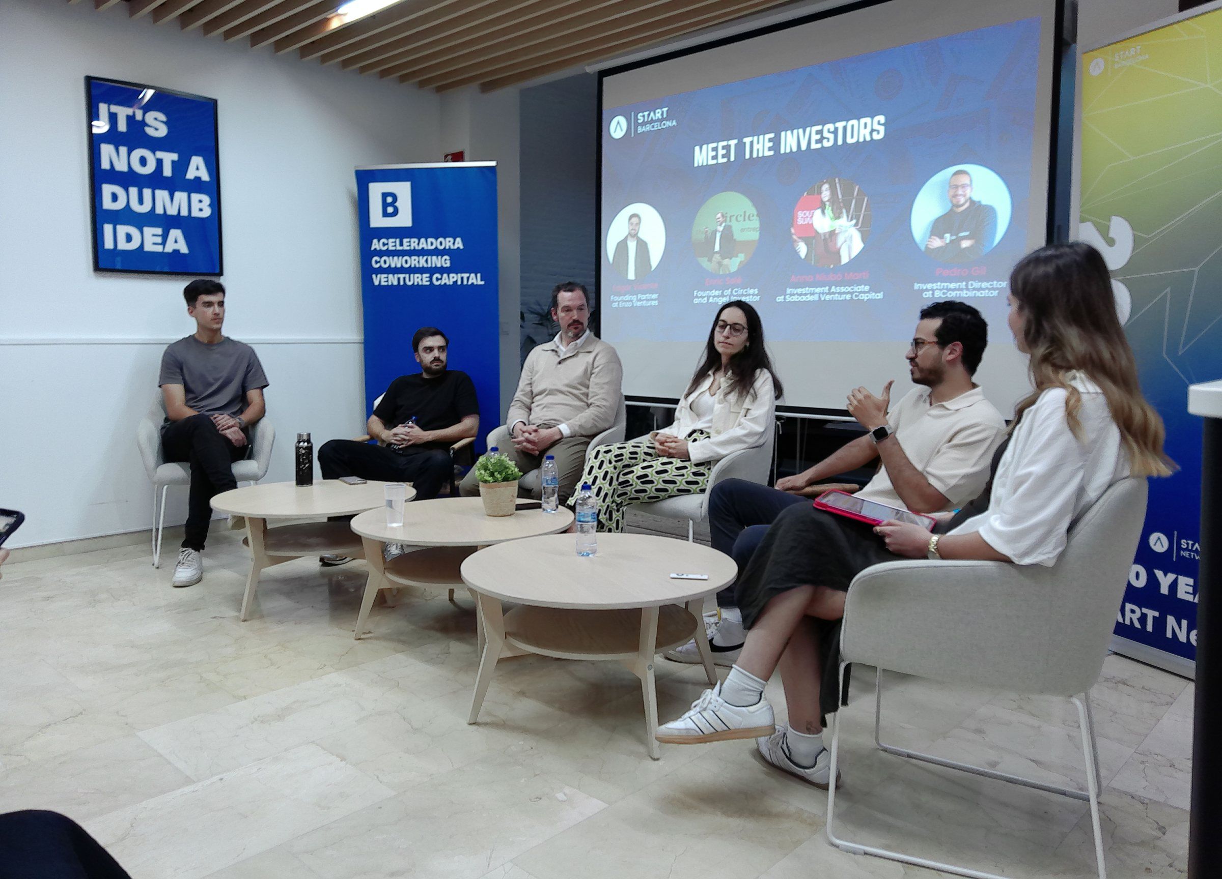 Four investors on a sofa panel under a 'Meet the investors' slide, a poster reading 'It's not a dumb idea' on the wall